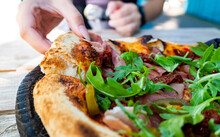 Woman Hand Takes A Slice Of Meat Neapolitan Pizza With Mozzarella Cheese, Ham, Bacon, Tomato, Spices And Jalapeno In Cafe