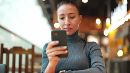 Portrait of beautiful young woman who uses mobile phone and looks in screen, reading text or texting and communicating on social networks. Girl sitting in public place, in shopping mall or restaurant.