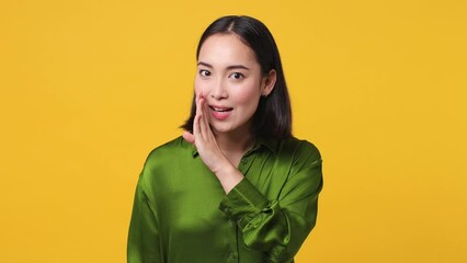 Wall Mural - Mysterious funny young woman of Asian ethnicity 20s she wear green shirt whispering gossip and tells secret behind her hand sharing news isolated on plain yellow color wall background studio portrait