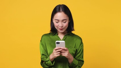 Wall Mural - Happy smiling calm young woman of Asian ethnicity 20s she wear green shirt hold use mobile cell phone typing browsing chatting send sms isolated on plain yellow color wall background studio portrait