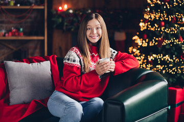 Poster - Portrait of cheerful lovely girl sitting couch hands hold coffee mug tree garland lights house indoors