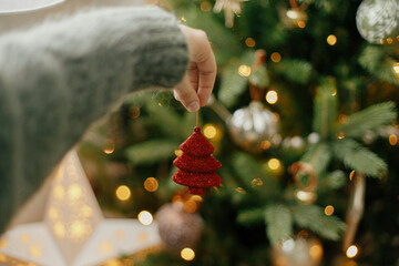 Hand holding red christmas tree on background of illuminated tree with golden lights. Woman decorating christmas tree and holding glitter ornament in hand in festive room. Atmospheric time