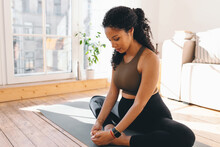 Side View Of Pregnant Woman Practicing On Floor, Siting On Mat In Butterfly Yoga Pose, Stretching Inner Part Of Thigh, Breathing Deeply, Wearing Leggings And Smart Watch To Check Pulse