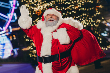 Poster - Portrait of positive cheerful grandfather santa carry presents bag arm hand waving congratulate tree garland lights evening outdoors