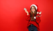 Satisfied Customer With A Credit Card And A Gift In His Hands On A Red Background. Young Woman In Santa Hat And Sweater, Christmas Sales.