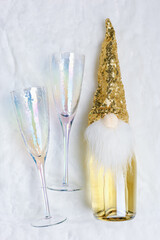 New Year celebrations aesthetic flat lay, champange bottle decorated shiny cap hat and beard from fun dwarf, two wine glasses on white fur background, holiday winter concept, alcogolic drinks