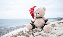 Bdsm Accessories On A Plush Christmas Bear In A Santa Claus Hat On The Background Of Rocks And The Sea