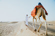 © oneinchpunch - Man wearing traditional clothes, taking a camel out on the desert sand, in Dubai