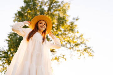 Wall Mural - low-angle portrait of smiling young woman in straw hat and white dress standing posing on beautiful 