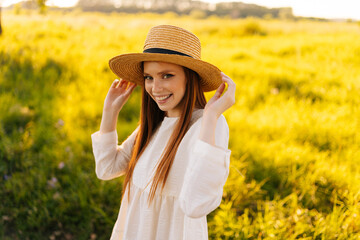 Wall Mural - Medium shot portrait of pretty positive ginger young woman wearing straw hat and white dress standing posing on beautiful field of green grass looking at camera, in sunset sunlight on summer evening.