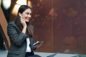 Side view of a business woman holding laptop near office outdoors, using mobile phone. Traveling, working outdoors in city, working on laptop computer, happy.