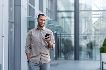 A young handsome Hispanic, African-American man walks down the street near an office center. He is holding a phone in his left hand, his right hand is in his pocket. Uses, reads the news, smiles.