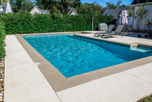 A Rectangular New Swimming Pool With Tan Concrete Edges In The Fenced Backyard Of A New Construction House