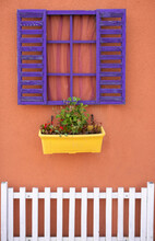 Colorful Geranium And White Fence Under Wooden Purple Window With Open Shutter On Orange Wall