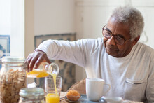 Portrait Of Senior Man Having Breakfast In Kitchen. Grey-haired Man In Glasses Calmly Sitting At Table Focused On Pouring Orange Juice Into Glass. Senior Peoples Life, Support And Health Care Concept