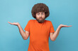 Clueless confused man with Afro hairstyle in T-shirt shrugging shoulders as doesn't know answer, can`t make decision, being uncertain, not sure. Indoor studio shot isolated on blue background.