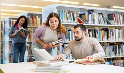 Canvas Print - Interested young bearded man working with his female colleague on joint project in public library, looking for information in books and Internet