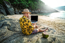 Man in the summer hat , a businessman, digital nomad working with laptop on the rocky beach.  Freelancer. Tropics.