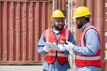 Two short black hair man with moustache and beard dressed in yellow hardhat, red safety vest and white protective glove working during the day under sunlight. There are container in the work area.