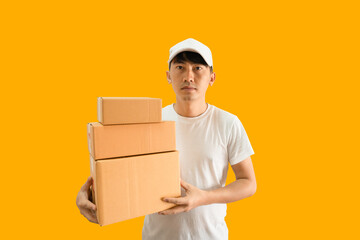 Young Asian delivery man wearing cap and white blank t-shirt holding parcel post box isolated on yellow background. express delivery service concept.