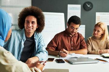 Poster - Happy young man with thick dark hair looking at Muslim female colleague in blue hijab during conversation by workplace