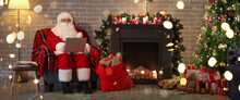 Santa Claus With Laptop Sitting In Armchair Near Fireplace On Christmas Eve