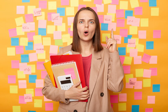 Indoor shot of inspired exited young woman student holding copybooks and paper folders, having idea about science project, posing against sticky notes for leaving reminder.