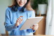 © Jirapong - Portrait of a teenage Asian woman using a tablet, wearing headphones to study online via video conferencing on a wooden desk in library