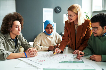 Wall Mural - Confident blond teacher in eyeglasses looking at one of students while explaining information in document at lesson of foreign language