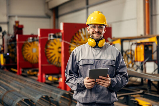A heavy industry worker is using tablet in factory while smiling a the camera.