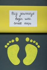Wall Mural - Card with phrase Big Journeys Begin With Small Steps and paper feet on dark background, top view. Motivational quote