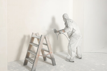 Wall Mural - Decorator in protective overalls painting wall with spray gun indoors