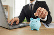 © Studio Romantic - Businessman puts money in his piggy bank. Man in a suit uses a modern laptop notebook PC and puts a coin in a blue piggy bank on his office desk. Business, finance, investment, online banking concept