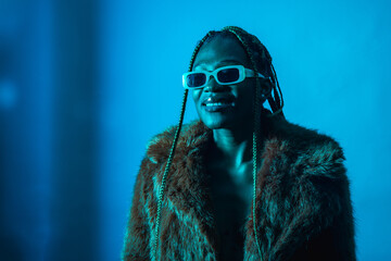 Wall Mural - Attractive black ethnic woman with braids with blue led lights, wearing a fur coat looking at the camera