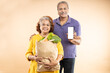 © GAJENDRRA BHATI  - Happy senior indian couple hold grocery paper bags with food and blank smart phone screen mockup isolated on beige background. studio shot.