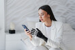 Beautiful brunette hispanic girl with artificial hand in white suit sitting on bed holds phone reads messages. Rehabilitation after trauma. Orthopaedics and hi tech medical care. Cybernetic medicine.