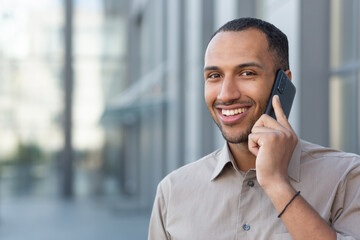 Close up photo. Handsome hispanic, african american man standing on the street and talking on the phone, waiting for an appointment, traveling. He looks at the camera, smiles