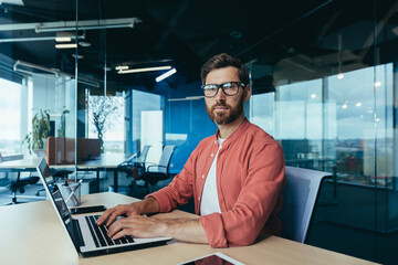 portrait of a successful programmer inside the office, a man with a beard, glasses and a red shirt l