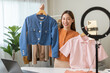 © KMPZZZ - vlogger, blogger small SME business asian beautiful woman using mobile phone video call for sell clothes live stream selling online, show product present detail at home office, entrepreneur ecommerce