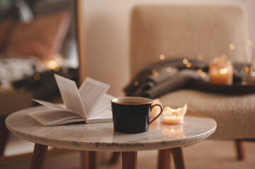 Cup of tea with paper open book and burning scented candles on marble table over cozy chair and glowing lights in bedroom closeup. Winter holiday season.