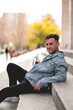 © Samuel Ponce - Young blonde model guy with sunglasses, denim jacket and black jeans sitting on stone stairs in a park in the city in the  afternoon