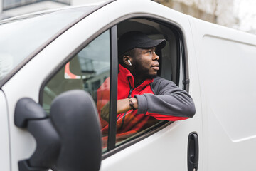 Wall Mural - Black young adult delivery guy wearing red pullover and black cap driving white van leaning outside the window to look behind. Horizontal shot. High quality photo