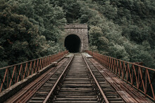 Rusty Train Tracks Lead Into A Dark Tunnel In A Desaturated Landscape