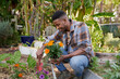 © StratfordProductions - Profile view of happy young black man holding flowers and spade by flowerbed in garden center
