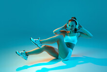 Young Sportive Girl In White Sportswear And Headphones Training, Doing Crunches Isolated Over Blue Studio Background In Neon Light