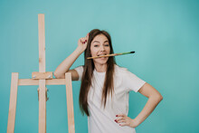 Young Girl In Casual Clothes With Long Hair, Holding A Brush In Her Mouth, Leaning On An Easel On A Turquoise Background. Mockup For Creative Ideas, Hobby, Arts, Leisure.