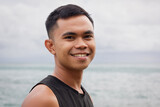 Fototapeta Sypialnia - Smiling portrait of young Filipino man by the sea on cloudy day. Asian male model concept