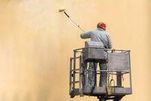 Man On A Lifting Platform Painting The Building Wall With A Roller Exterior Outdoors. Worker On A Ladder Manually Painting Yellow Wall On Construction Site