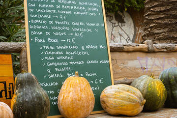 Horizontal still life of four horizontal pumpkins, two orange and two green, on a wooden bench. In the background an old blackboard, written with white chalk the menu in Spanish of a street restaurant