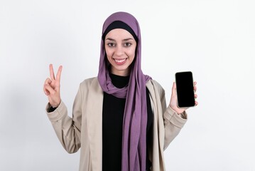 young beautiful muslim woman wearing hijab and jacket over white background holding modern device showing v-sign